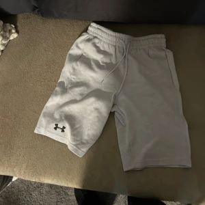 Grå shorts från Under Armour - Säljer ett par grå shorts från Under Armour med elastisk midja och dragsko. Perfekta för träning eller en avslappnad dag. Under Armour-loggan finns på benet.