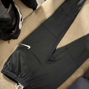 Svarta träningsbyxor från Nike - Säljer ett par svarta träningsbyxor från Nike med Dri-FIT-teknologi. Byxorna har en vit Nike-logga på benet och dragsko i midjan för justerbar passform. Perfekta för träning och sportaktiviteter.