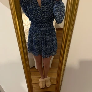 Blå mönstrad klänning från Zara - Säljer en blå mönstrad klänning med långa ärmar och v-ringad hals. Klänningen har en smockad midja som ger en fin passform och volangdetaljer vid fållen. Perfekt för en stilfull look. Klänningen är från Zara och i fint skick🥰🥰