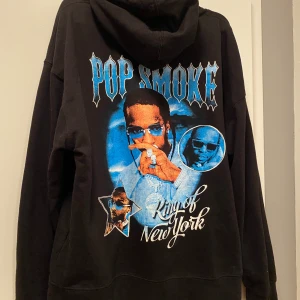 Svart Pop Smoke hoodie strl m - Säljer en svart hoodie med Pop Smoke-tryck på baksidan. Texten 'King of New York' och bilder av artisten i blått och vitt ger en cool look. Perfekt för fans av Pop Smoke. Hoodien har en klassisk passform med känguruficka och dragsko i huvan.