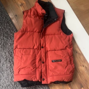 Röd dunväst från Canada Goose - Säljer en snygg röd dunväst från Canada Goose. Västen har en klassisk design med knappar och två stora fickor framtill. Perfekt för kyliga dagar när du vill hålla värmen utan att kompromissa med stilen.