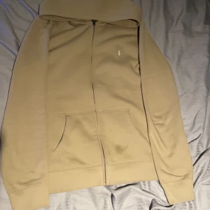 Ralph Lauren Zip Hoodie Beige - Köpt på kidsbtanfstore. Säljer en stilren beige hoodie från Ralph Lauren med dragkedja och fickor framtill. Den har en broderad logotyp på bröstet och är perfekt för en avslappnad look. Passar bra till både jeans och joggers. 