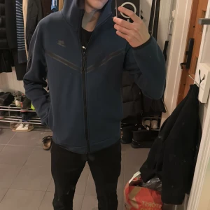 Blå Nike tech fleece  - Säljer en blå Nike tech med dragkedja och huva. Tröjan har en ficka med dragkedja på ärmen och är perfekt för kyligare dagar. Den har en sportig look och är bekväm att bära. Har mitt namn inskrivet på insida lapp.