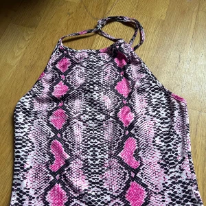 Rosa och svart ormmönstrad halterneck topp - Snygg halterneck topp med ormmönster i rosa och svart. Perfekt för en trendig look. Toppen har en knytning i nacken och är ärmlös, vilket ger en stilren och modern känsla.