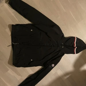 Svart windbreaker från Moncler - Snygg svart jacka från Moncler med huva och dragkedja. Jackan har en broderad logga på ärmen och detaljer i rött och vitt på huvan. Perfekt för en stilren look. Se defekter på bild 3 och 4. Pris kan diskuteras✌️