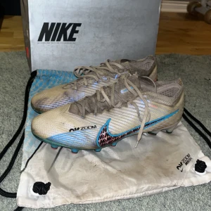 Nike fotbollsskor med snörning - Snygga Nike fotbollsskor med snörning och en modern design. Skorna har dobbar för både konstgräs och naturligt gräs. Skorna har  en vit bas med blå och rosa detaljer samt en slitstark sula med dobbar för optimalt grepp på planen. Perfekta för fotbollsspelare som vill ha både stil och funktion. Skick 7/10. Skriv vid minsta fundering.