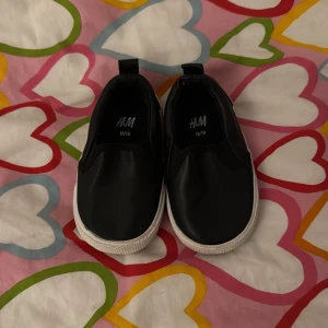 Svarta slip-on skor från H&M - Säljer ett par svarta slip-on skor från H&M i storlek 18/19. Skorna har en vit sula och är perfekta för små fötter. De är lätta att ta på och av tack vare den elastiska designen.