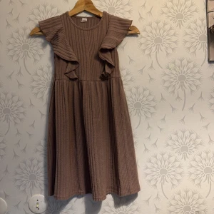 Brun klänning med volangdetaljer - Charmig brun klänning med volangdetaljer på axlarna. Den har en ribbad struktur och är ärmlös, vilket ger en luftig och bekväm känsla. Perfekt för en avslappnad dag ute eller en mysig kväll hemma.