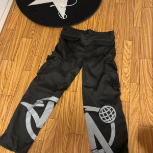 Svarta cargopants med tryck - Coola svarta cargopants med grått tryck och flera fickor. Perfekta för en avslappnad stil med en touch av streetwear. Byxorna har en loose passform och är tillverkade i ett slitstarkt material. En av dom som ska hålla i bältet har lossnat