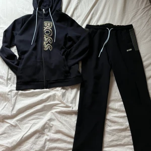 Sällsynt tracksuit från Boss i marinblå med stor logga - Snygg sällsynt marinblå tracksuit från Boss med dragkedja och stor logga framtill. Den har en justerbar huva med snören och ribbade muddar. Perfekt för en avslappnad stil. Nypris 3298:- SEK 