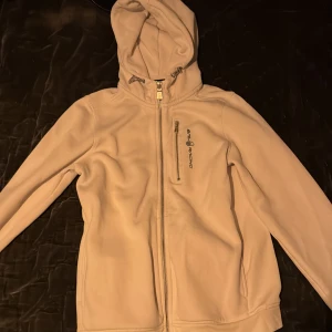 Beige fleecejacka från Sail Racing - Säljer en beige fleecejacka från Sail Racing med huva och dragkedja framtill. Jackan har en enkel design med en liten logotyp på bröstet. Perfekt för kyliga dagar.