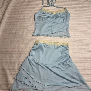 Ljusblå tvådelad klänning med spetsdetaljer - Charmig ljusblå tvådelat set med en topp som har knytband och spetsdetaljer vid bysten. Kjolen har matchande spets i midjan och har en asymmetrisk design. Perfekt för en somrig, ”mermaid” look!🤍🏖️
