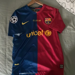 Barcelona tröja Messi 2009 - Säljer en ikonisk Barcelona fotbollströja från Nike i blått och rött. Tröjan har korta ärmar och är prydd med klubbens emblem och Champions League-märke. Den har även texten 'més que un club' och spelarens namn och nummer på baksidan.