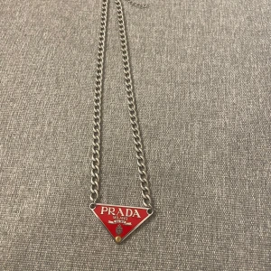 Halsband från Prada med röd triangel - Snyggt halsband från Prada med en röd triangelformad platta som har märkets logotyp. Kedjan är i silverfärg och ger en stilren look. Perfekt för att ge en touch av lyx till din outfit.