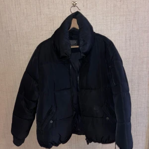 Svart pufferjacka - Säljer en svart pufferjacka med dragkedja och hög krage. Jackan har långa ärmar och två fickor framtill med knappar. 