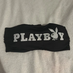 Svart bandeau bikini från Playboy - Trendig svart bandeau bikiniöverdel från Playboy med stort tryck fram och små kaninmotiv i vitt och rosa på baksidan. Den har en knytning bak för justerbar passform. Perfekt för en stilren look vid poolen eller stranden.