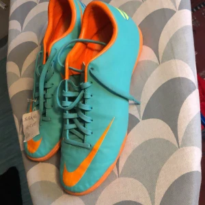 Turkosa och orange Nike fotbollsskor - Snygga turkosa fotbollsskor från Nike med orange detaljer och snörning. Perfekta för fotbollsplanen med en iögonfallande design.