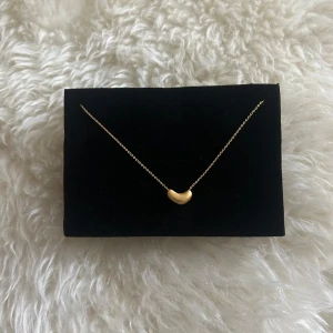 Guldhalsband med böjd berlock - Elegant guldhalsband med en unik böjd berlock. Kedjan är tunn och smidig, vilket ger ett stilrent intryck. Perfekt för att lägga till en touch av elegans till din outfit. Märkt med 'Muli' vid låset.
