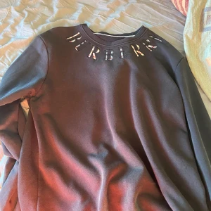 Svart tröja från Burberry - Säljer en stilren svart tröja från Burberry med märkets namn broderat i ett mönster på framsidan. Tröjan har en klassisk design med rund hals och långa ärmar. Perfekt för en avslappnad men ändå elegant look. Säljer den snabbt och billigt då den bara tar plats i garderoben.