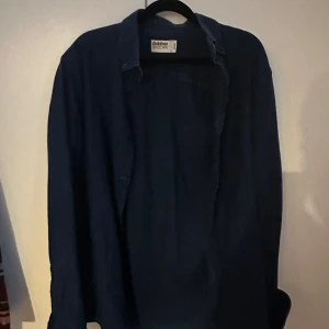 Mörkblå jeansskjorta från Dobber - Snygg mörkblå jeansskjorta från Dobber. Perfekt för en stilren look med klassiska knappar framtill. Skjortan har en avslappnad passform och är tillverkad i ett mjukt material.