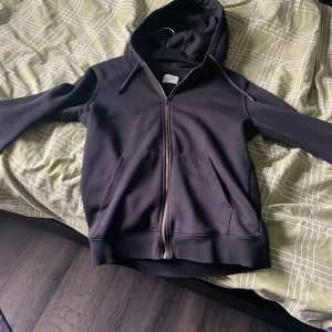 Svart hoodie med dragkedja - Säljer en stilren svart hoodie med dragkedja och fickor framtill. Perfekt för en avslappnad look. Den har en klassisk passform och är tillverkad i ett mjukt material som gör den bekväm att bära.