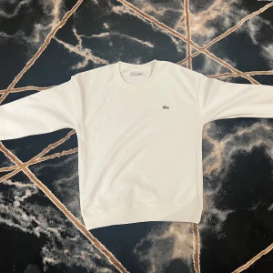 Vit sweatshirt från Lacoste - Säljer en stilren vit sweatshirt från Lacoste med det klassiska krokodilmärket på bröstet. Tröjan har långa ärmar och en rund halsringning. Perfekt för en avslappnad och snygg look.