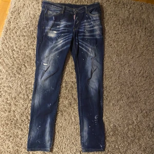 Blå jeans från Dsquared2 - Snygga blå jeans från Dsquared2 med coola slitningar och färgstänk för en unik look. De har en klassisk femficksdesign och en knappgylf. Perfekta för dig som vill ha en edgy stil.