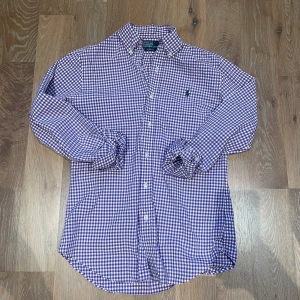 Lila rutig skjorta från Ralph Lauren - Snygg lila och vit rutig skjorta från Ralph Lauren med broderad logga på bröstet. Skjortan har en klassisk krage och knappar framtill. Perfekt för en stilren look. Skjortan har storlek S och passar 165-178 Modellen är 170cm och 65kg. Skriv vid minsta fundering!