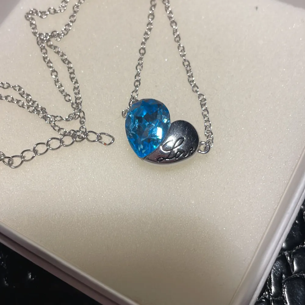 Elegant halsband med en silverkedja och ett hängsmycke i form av ett hjärta. Ena halvan av hjärtat är en klar blå sten, medan den andra halvan är i silver med ingraverad text. . Asusteet.