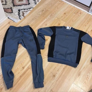 Blå träningsset från Adidas - Säljer ett snyggt blått träningsset från Adidas med svarta detaljer. Tröjan har långa ärmar och byxorna har en bekväm resår i midjan. Perfekt för träning eller en avslappnad dag. Adidas-loggan syns både på tröjan och byxorna. TRÖJA XS BYXOR S