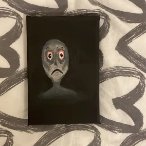 Målning av ansikte med sorgsna ögon - En unik målning på en svart bakgrund som visar ett ansikte med stora, sorgsna ögon och en nedåtvänd mun. Ansiktet är målat i grå toner med röda detaljer runt ögonen, vilket ger en känsla av djup och uttryck. Som vissar även känslan av besvikelse 