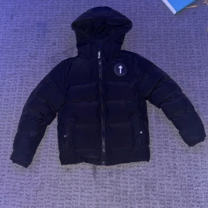 Trapstar jacka - Säljer en svart pufferjacka från Trapstar med huva och dragkedja. Jackan har ett tryck med texten 'It's a Secret' på bröstet och 'Trapstar' på ryggen. Perfekt för kyliga dagar.