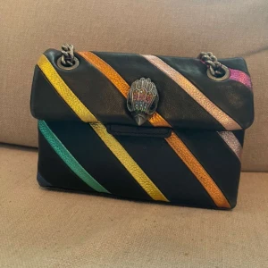 Kurt Geiger väska - Snygg axelväska i svart skinn med diagonala ränder i regnbågens färger. Väskan har en kedjerem och ett dekorativt spänne framtill. Perfekt för att ge din outfit en färgglad touch! Tappat lite färg och lite sliten (bild 4) annars fin