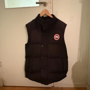 Svart dunväst från Canada Goose - Säljer en svart dunväst från Canada Goose med knappar och ett märke på bröstet. Västen är perfekt för kyliga dagar och har en klassisk design som passar till det mesta. Den är tillverkad i Kanada och har en hög krage för extra värme.