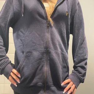 Mörkblå hoodie från Lyle & Scott - Säljer en snygg mörkblå hoodie från Lyle & Scott med dragkedja och huva. Den har en liten gul logga på bröstet och praktiska fickor framtill. Perfekt för en avslappnad stil. 