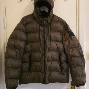 Brun pufferjacka från Replay - Säljer en brun pufferjacka från Replay med en cool logga på ryggen. Jackan har en dragkedja framtill och en huva med dragsko. Insidan är fodrad i rött för en snygg kontrast. Perfekt för kyliga dagar!