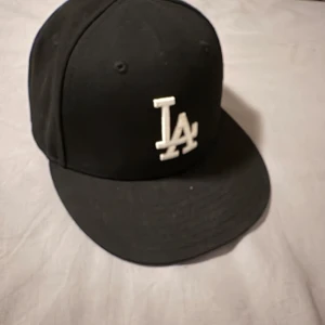 Svart keps från New Era - Snygg svart keps från New Era med LA-logga i vitt framtill. Skick 7/10 55.8 cm