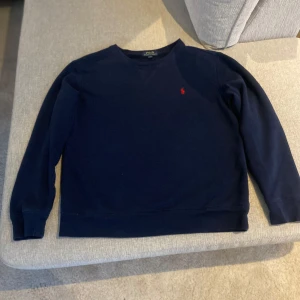 Mörkblå tröja från Ralph Lauren - Säljer en stilren mörkblå tröja från Ralph Lauren med det klassiska röda logotypbroderiet på bröstet. Tröjan har långa ärmar och en rund halsringning. Perfekt för en avslappnad och snygg look. Det står att det är L men den passar M och S också!