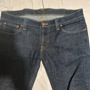 Blå jeans från Nudie Jeans - Snygga blå jeans från Nudie Jeans med klassisk femficksdesign och kontrasterande sömmar. De har en knappgylf och en läderpatch med logotyp baktill. I Slim fit. Storlek 32 32. Perfekta för en avslappnad stil.