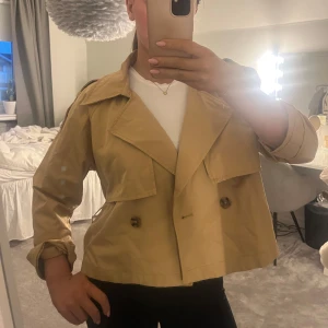 Beige croppad trenchcoat - Snygg beige croppad trenchcoat med dubbelknäppning och klassisk krage. Perfekt för vår och höst. Jackan har långa ärmar och en stilren design som passar till många olika outfits. Skärp finns! 