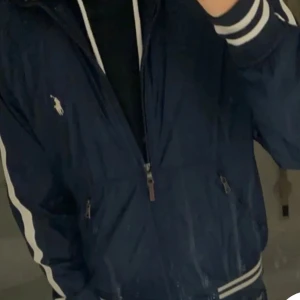 Mörkblå jacka från Ralph Lauren - Säljer en snygg mörkblå jacka från Ralph Lauren med dragkedja och ribbade muddar. Jackan har ett broderat logotypmärke på bröstet och vita detaljer längs ärmarna. Perfekt för en stilren look.