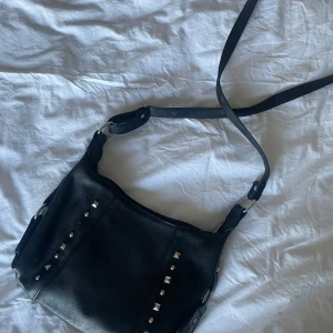 Nitväska - Snygg svart axelväska i skinn med nitar på framsidan. Väskan har en justerbar axelrem och en praktisk dragkedja på baksidan för extra förvaring. En nit har lossnat, annars i fint skick