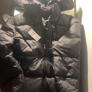 Svart dunjacka från Polo Ralph Lauren - Säljer en snygg svart dunjacka från Polo Ralph Lauren. Jackan har en dragkedja framtill och en liten broderad logga på bröstet. Perfekt för kyliga dagar med sin varma och puffiga design. Den är för stor på mig det är därför jag säljer den.
