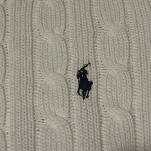Vit cable knit från Ralph Lauren - Säljer en klassisk vit stickad tröja från Ralph Lauren med kabelstickat mönster och den ikoniska logon på bröstet. Perfekt för en stilren look.