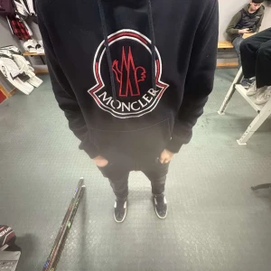 Svart hoodie från Moncler - Säljer en snygg svart hoodie från Moncler med stor logga på bröstet. Tröjan har långa ärmar och en praktisk magficka. Perfekt för kyliga dagar eller som en stilren vardagslook.