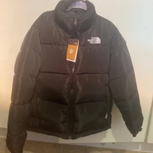 Svart pufferjacka från The North Face - Säljer en svart pufferjacka från The North Face med hög krage och dragkedja framtill. Jackan har lång ärm och klassisk logga på bröstet. Perfekt för kyliga dagar och riktigt snygg till streetwear-stilen.