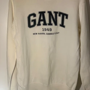 Vit stickad tröja från GANT - Säljer en stilren vit tröja från GANT med texten 'GANT 1949 New Haven, Connecticut' på framsidan. Tröjan har långa ärmar och ribbade muddar vid ärmslut och nederkant. Perfekt för en avslappnad stil.