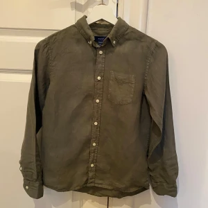 Olivgrön skjorta från GANT - Säljer en stilren olivgrön skjorta från GANT i storlek 146/152 cm. Skjortan har långa ärmar och en klassisk krage med knappar framtill. Perfekt för en avslappnad men ändå snygg look.