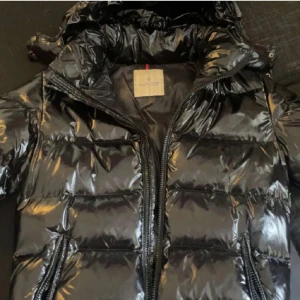 Svart glansig dunjacka från Moncler - Säljer en svart glansig dunjacka från Moncler med dragkedja och huva. Jackan har en quiltad design och Moncler-logga på ärmen. Perfekt för kyliga dagar med stil. (Har en defekt därav priset)