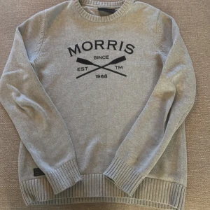 Grå tröja från Morris R. Club - Säljer en stilren grå tröja från Morris R. Club med svart text på framsidan. Tröjan har långa ärmar och ribbade muddar vid ärmslut och nederkant. Perfekt för en avslappnad och grisch stil. Nypris 1400kr